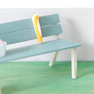 Könnte beinhalten: Eine Miniatur-Parkszene mit einer tealfarbenen Bank, einem orangefarbenen Objekt und einem kleinen Eimer mit Eis. Ein Fußball liegt auf dem Boden, und eine kleine Figur steht auf einem grauen Würfel. Die Szene befindet sich auf einer grünen Oberfläche.