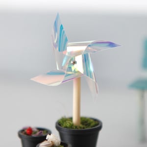 Könnte beinhalten: Ein Miniatur-Windrad mit irisierender Oberfläche auf einem Holzstab, eingepflanzt in einen kleinen schwarzen Topf mit grünem Moos. Zwei weitere kleine schwarze Töpfe mit Moos, winzigen Muscheln und einer roten Perle stehen daneben.