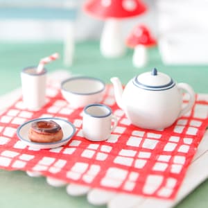 Könnte beinhalten: Miniatur-Picknick-Szene mit einer rot-weiß karierten Decke, einer Teekanne, einer Tasse, einer Schüssel und einem Teller mit Gebäck. Eine kleine schwarze Laterne mit gelbem Schein steht daneben. Eine blaue Bank und Pilzdekorationen befinden sich im Hintergrund.