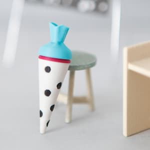 Könnte beinhalten: Eine Miniatur-Schultüte in Weiß mit schwarzen Punkten und einem hellblauen Verschluss, platziert neben einem kleinen grünen Hocker. Im Hintergrund steht ein Miniatur-Schreibtisch aus Holz mit weißem Papier und einem Schild mit der Aufschrift Happy Birthday.