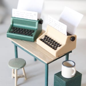 Könnte beinhalten: Eine Miniaturszene mit einer grünen Schreibmaschine und einer beigen Schreibmaschine, in denen jeweils ein weißes Blatt Papier steckt, platziert auf einem kleinen rechteckigen Tisch mit passendem Hocker und einer weißen Tasse auf einem grünen Sockel.