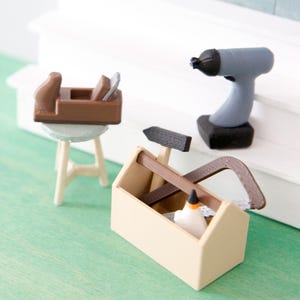 Può includere: Set di piccoli attrezzi da falegname su una superficie verde. Include una cassetta degli attrezzi, una sega, un martello, un trapano e una piccola pialla in legno su uno sgabello. Gli attrezzi sono nei toni del marrone, beige, grigio e nero.