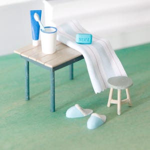 Könnte beinhalten: Miniatur-Badezimmer-Szene mit einem kleinen Holztisch, einer blauen Tube Zahnpasta, einer Zahnbürste in einem weißen Becher und einem Seifenstück mit dem Wort "SEIFE". Ein gestreiftes Handtuch, ein kleiner Hocker und blaue Hausschuhe vervollständigen die Szene.