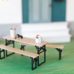 Könnte beinhalten: Miniatur-Picknicktisch mit Bänken, mit hellbrauner Tischplatte und schwarzen Rahmen. Auf dem Tisch stehen ein kleiner Becher mit Strohhalm, eine Sonnenbrille und ein Eisbehälter. Eine Lichterkette mit bunten Lichtern hängt darüber.