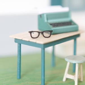 Könnte beinhalten: Eine Miniatur-Schreibmaschine in Blaugrün und eine Brille mit schwarzem Rand stehen auf einem hellen Holztisch mit blaugrünen Beinen. Neben dem Tisch befindet sich ein kleiner, runder, heller Hocker mit weißen Beinen für Miniatur-Szenen.