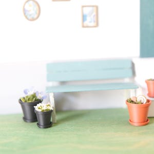 Op de afbeelding: Miniatuurscène met een lichtblauwe bank, kleine terracotta potten en zwarte potten met planten. De scène is geplaatst tegen een witte muur met ingelijste kunstwerken, wat een grillige, decoratieve weergave creëert. De grond is een groen oppervlak.