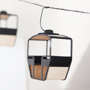 Könnte beinhalten: Zwei Miniatur-Seilbahngondeln, die an einer dunklen Schnur hängen. Jede Gondel verfügt über einen schwarzen Metallrahmen, eine Basis in hellem Holzton und ein offenes Design mit gewölbtem Dach, aufgehängt an einem dünnen schwarzen Haken.
