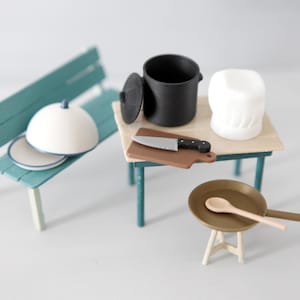 Puede incluir: Escena de cocina en miniatura con un banco turquesa que sostiene una campana blanca y platos. Una pequeña mesa con una olla negra, gorro de chef, tabla de cortar con cuchillo y una sartén con una cuchara. Un juego de cocina en miniatura.