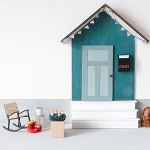 May include: A miniature wooden house facade with a teal wall, a light blue door, and a black mailbox labeled POST. The structure features white steps, a roof with pastel bunting, a rocking chair, red boots, a small stool, and a green wreath.