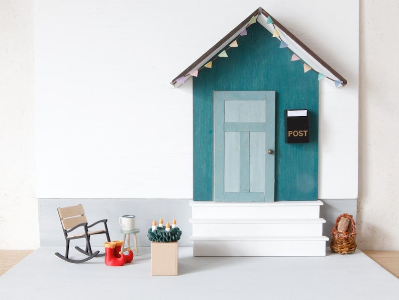 May include: A miniature wooden house facade with a teal wall, a light blue door, and a black mailbox labeled POST. The structure features white steps, a roof with pastel bunting, a rocking chair, red boots, a small stool, and a green wreath.