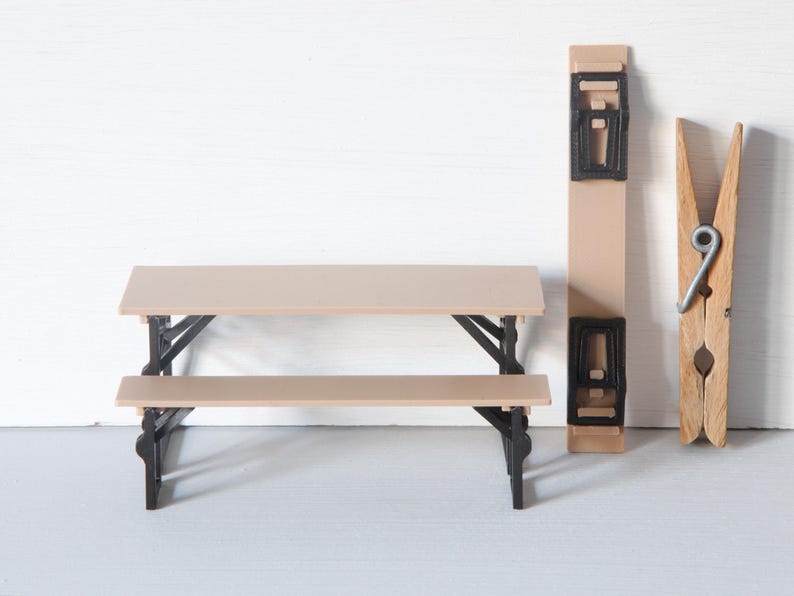 May include: A miniature picnic table with a tan tabletop and bench, supported by black legs. A matching tan and black object and a large wooden clothespin are also present, all against a white background.