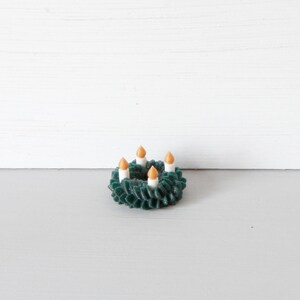 May include: A miniature dark green wreath featuring four small white candles with orange flames. The wreath has a textured, layered appearance and is circular in shape, designed as a small decorative item with a festive, seasonal aesthetic.