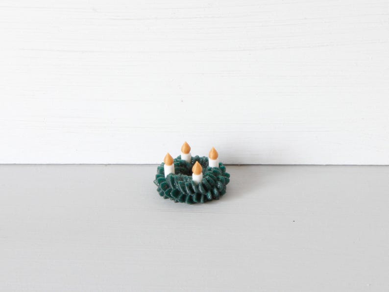 May include: A miniature dark green wreath featuring four small white candles with orange flames. The wreath has a textured, layered appearance and is circular in shape, designed as a small decorative item with a festive, seasonal aesthetic.