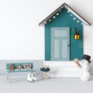 May include: A miniature house facade with a teal wall, a light blue door, and a brown roof with colorful bunting. A teal bench holds gingerbread cookies and white ice skates. A snowman with a black top hat and a broom stands near a small table.