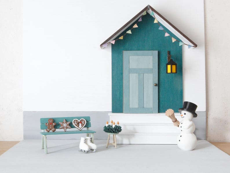 May include: A miniature house facade with a teal wall, a light blue door, and a brown roof with colorful bunting. A teal bench holds gingerbread cookies and white ice skates. A snowman with a black top hat and a broom stands near a small table.