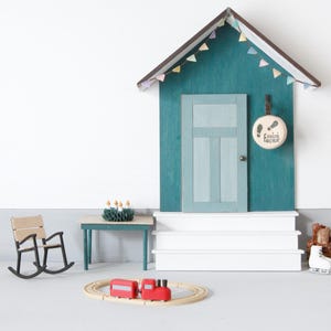 May include: A miniature wooden house facade with a teal wall, a light blue door, and white steps. A round sign with footprints and the text "mini factur" hangs by the door. Includes a small rocking chair, a table with a wreath, and a toy train.