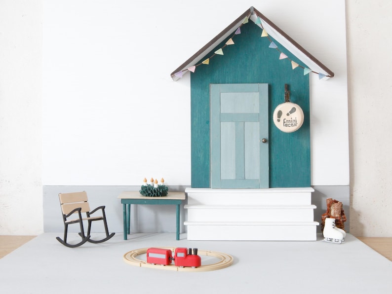 May include: A miniature wooden house facade with a teal wall, a light blue door, and white steps. A round sign with footprints and the text "mini factur" hangs by the door. Includes a small rocking chair, a table with a wreath, and a toy train.