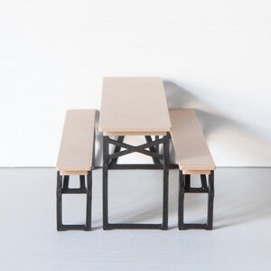 May include: A miniature picnic table set with a light wood tabletop and benches. The table and benches have black metal frames. The set is designed for a dollhouse or miniature scene.