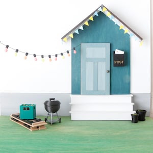 May include: A miniature outdoor scene featuring a teal house with a door, steps, and a black mailbox labeled "POST." A string of colorful lights hangs above. Includes a grill, a green crate, and small potted plants.