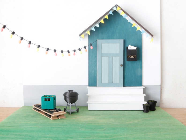May include: A miniature outdoor scene featuring a teal house with a door, steps, and a black mailbox labeled "POST." A string of colorful lights hangs above. Includes a grill, a green crate, and small potted plants.