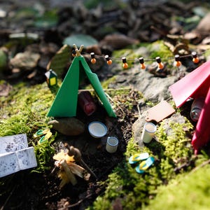 Może przedstawiać: Miniaturowa scena kempingowa z zielonym i czerwonym namiotem, łańcuchem świetlnym i akcesoriami kempingowymi. Scena obejmuje małą książkę, ognisko i maleńkie klapki, wszystko na posłaniu z mchu i liści.