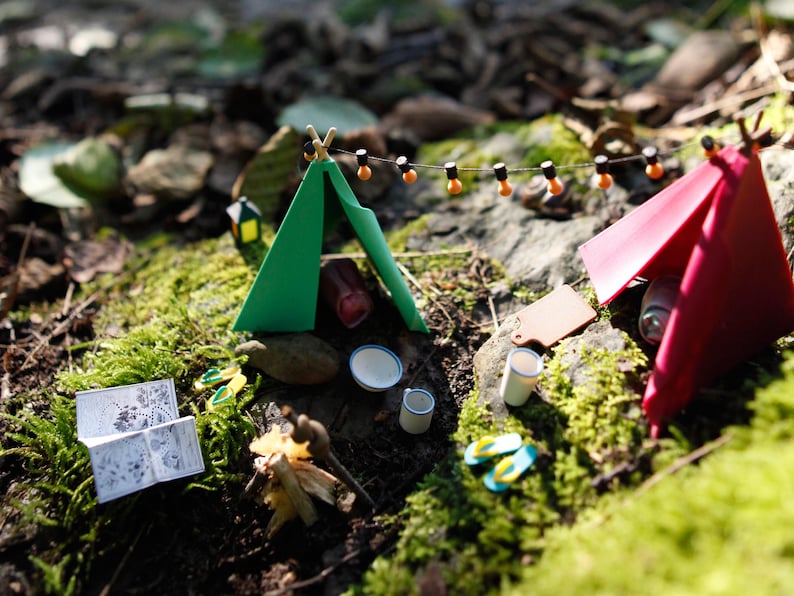 Może przedstawiać: Miniaturowa scena kempingowa z zielonym i czerwonym namiotem, łańcuchem świetlnym i akcesoriami kempingowymi. Scena obejmuje małą książkę, ognisko i maleńkie klapki, wszystko na posłaniu z mchu i liści.