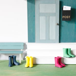 Könnte beinhalten: Eine Miniatur-Puppenhausszene mit einer hellblauen Tür, einem schwarzen Briefkasten mit der Aufschrift POST, einer hellblauen Bank und vier Paaren winziger Gummistiefel in Marineblau, Gelb, Pink und Grün auf weißen Stufen und grünem Boden.