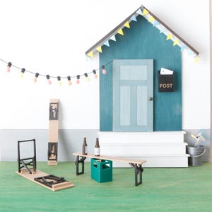 May include: A miniature outdoor scene with a teal house, white steps, and a green lawn. A string of colorful lights hangs above a wooden picnic table with two beer bottles. A folding chair and a tall, folded table are also present.