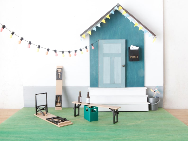 May include: A miniature outdoor scene with a teal house, white steps, and a green lawn. A string of colorful lights hangs above a wooden picnic table with two beer bottles. A folding chair and a tall, folded table are also present.