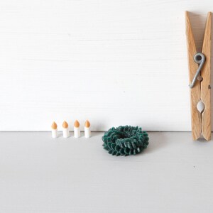 May include: A wooden clothespin, a small dark green circular wreath-like object with a textured surface, and four miniature white candles with orange tips arranged in a horizontal row on a flat surface.