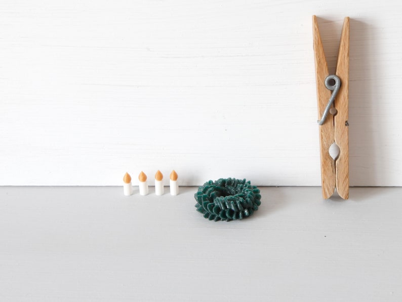May include: A wooden clothespin, a small dark green circular wreath-like object with a textured surface, and four miniature white candles with orange tips arranged in a horizontal row on a flat surface.