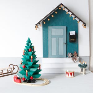 May include: A miniature holiday scene featuring a teal wooden house facade with a light-strung roof, a central door, and a black mailbox labeled POST. A green geometric Christmas tree with red ornaments and candles sits beside a wooden sled and train.