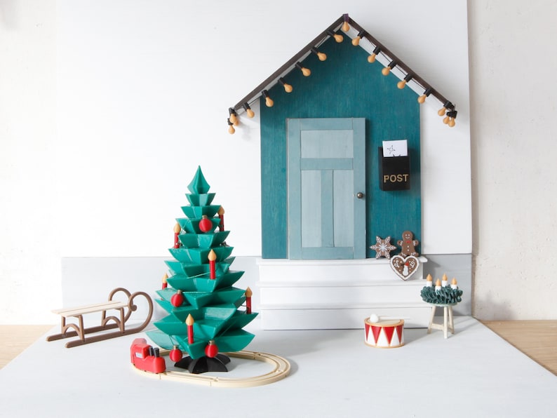 May include: A miniature holiday scene featuring a teal wooden house facade with a light-strung roof, a central door, and a black mailbox labeled POST. A green geometric Christmas tree with red ornaments and candles sits beside a wooden sled and train.