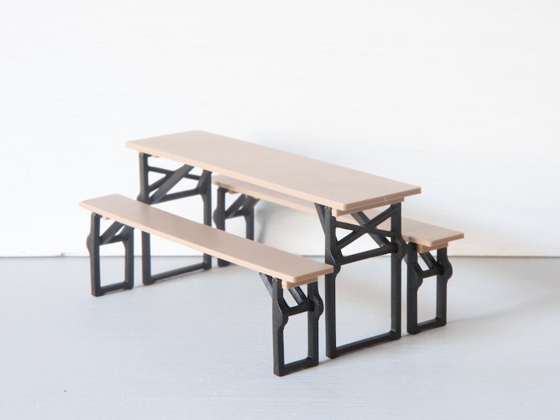 May include: A miniature picnic table with attached benches. The tabletop and benches are light brown, supported by black, angled legs. The table measures approximately 12 inches long, 6 inches wide, and 5 inches high.