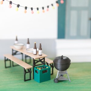May include: Miniature outdoor scene with a picnic table, benches, and a grill. The table holds bottles and a bowl. A green crate contains bottles. A string of lights hangs above the scene. The grill is black and silver.