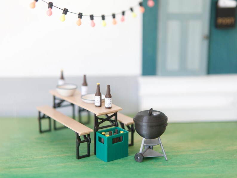May include: Miniature outdoor scene with a picnic table, benches, and a grill. The table holds bottles and a bowl. A green crate contains bottles. A string of lights hangs above the scene. The grill is black and silver.