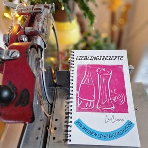 Könnte beinhalten: Ein spiralgebundenes Kochbuch mit dem Titel "LIEBLINGSREZEPTE" mit einem pinkfarbenen Einband mit weißen Illustrationen von Flaschen und einer Tomate. Der Text "La Cucina" und "VON MEINEN LIEBLINGSMENSCHEN" sind ebenfalls sichtbar. Das Kochbuch liegt auf einer Metalloberfläche.