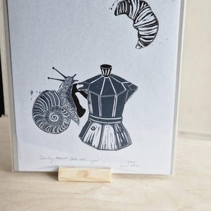 Puede incluir: Grabado en linóleo en blanco y negro que representa un caracol, una cafetera y un cruasán. El caracol está al lado de la cafetera y el cruasán está arriba. El texto "Snaily towards the late night" está en la parte inferior.