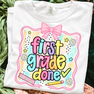 Könnte beinhalten: Weißes T-Shirt mit einem farbenfrohen Aufdruck "first grade done". Das Design umfasst eine rosa Schleife, Sterne, Gänseblümchen, ein Lineal und einen Bleistift. Der Text ist mehrfarbig, mit einem Häkchen neben dem Wort "done".