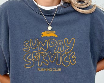 May include: Denim blue t-shirt with the text "SUNDAY SERVICE RUNNING CLUB" in yellow. The shirt has a relaxed fit and short sleeves. The text includes a graphic of a running rabbit.