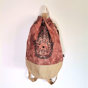 Op de afbeelding: Een rugzak met trekkoord en een rood-bruin tie-dye patroon met een zwart Hamsa hand ontwerp. De onderkant is van natuurlijk jute, met bijpassende schouderbanden.