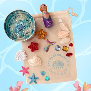 May include: A sensory play kit featuring a round metal tin filled with blue marbled dough and a small crab figure, a wooden mermaid peg doll, a beige drawstring bag with the text THE PLAYDOUGH TIN LET'S CREATE, resin stars, shells, and crystals.