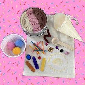 May include: A craft kit featuring a round metal tin labelled The Playdough Tin Let's Create containing multi-coloured dough, a white fabric drawstring bag with the same text, a white paper cone, a small wooden scoop, pom-poms, gems, and sticks.