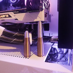 May include: Two metallic bullet-shaped objects, one bronze and one silver, stand upright on a white surface. The silver bullet has a textured design. The background includes computer components and wiring, with a purple hue.