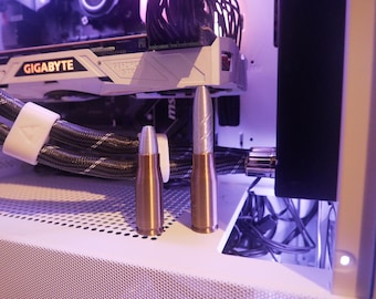 May include: Two metallic bullet-shaped objects, one bronze and one silver, stand upright on a white surface. The silver bullet has a textured design. The background includes computer components and wiring, with a purple hue.