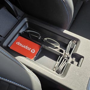 Könnte beinhalten: Mittelkonsole eines Autos mit einer roten Karte mit der Aufschrift "zapmapz" und einer Brille. Ein USB-Kabel und ein Ladeanschluss sind ebenfalls sichtbar. Die Konsole ist schwarz und grau.