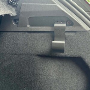 Könnte beinhalten: Ein schwarzer Kunststoffhaken, der an der inneren Seitenverkleidung eines Fahrzeugkofferraums montiert ist. Der Haken weist eine deutlich eingeprägte R5-Markierung auf seiner Oberfläche auf und dient als sicherer Befestigungspunkt für Gegenstände.