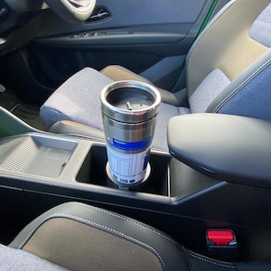 May include: A stainless steel travel mug with a blue and white graphic of a droid character, sitting in a car's cup holder. The mug has a black lid and a silver body. The car interior features gray and black seats.