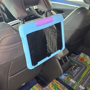 May include: A blue and purple tablet holder is attached to the headrest of a car seat. The tablet screen is black. The car interior includes gray seats and a colorful road map play mat.