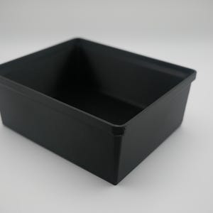 May include: A black rectangular storage bin with a slightly tapered design. The bin is empty and sits on a white surface. The sides are straight and the corners are squared.
